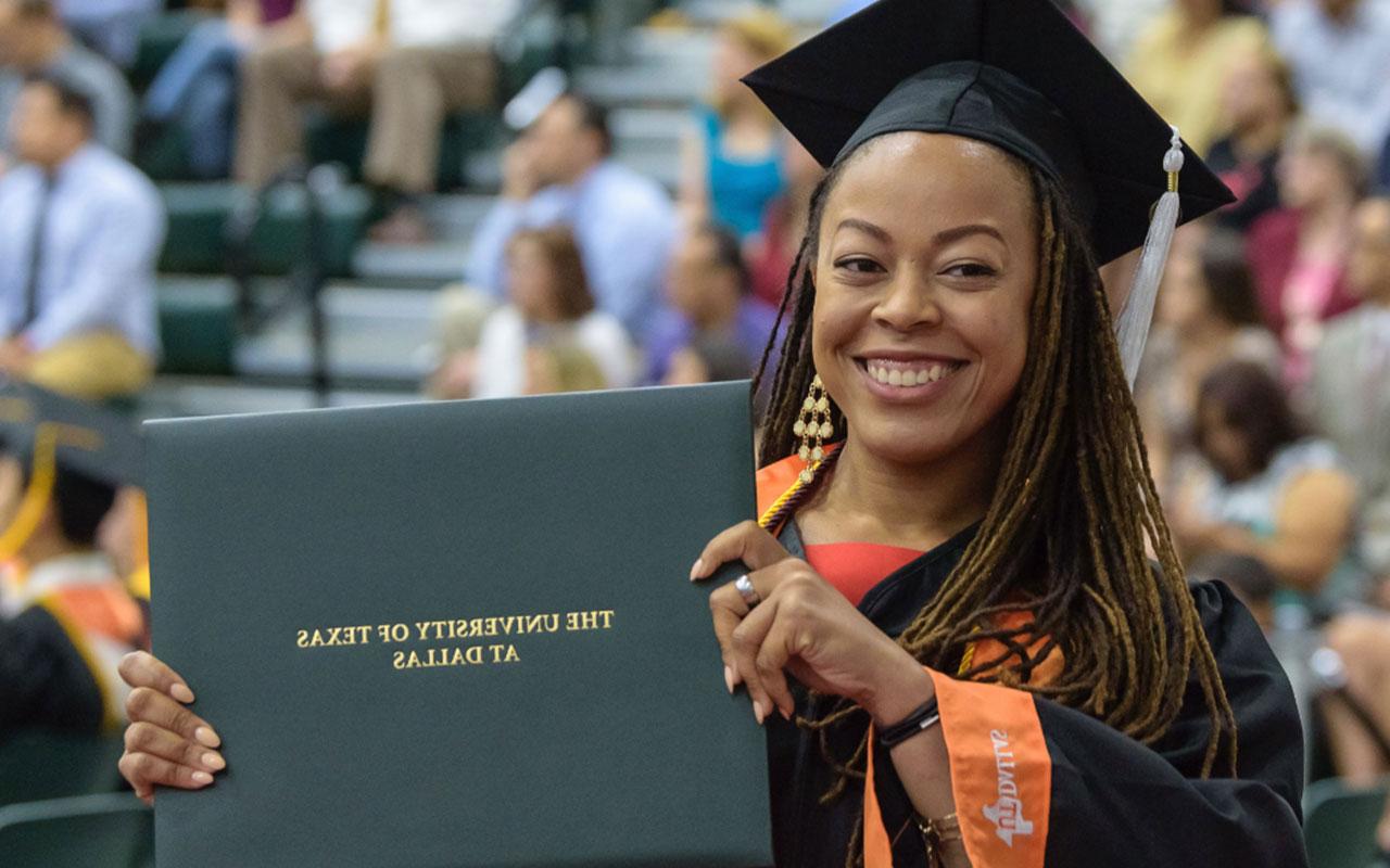 A graduate smiles while holding a diploma holder during commencement.